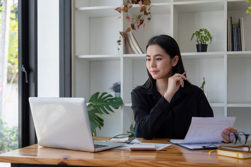 A young woman looking over at a laptop while holding a sheet of paper