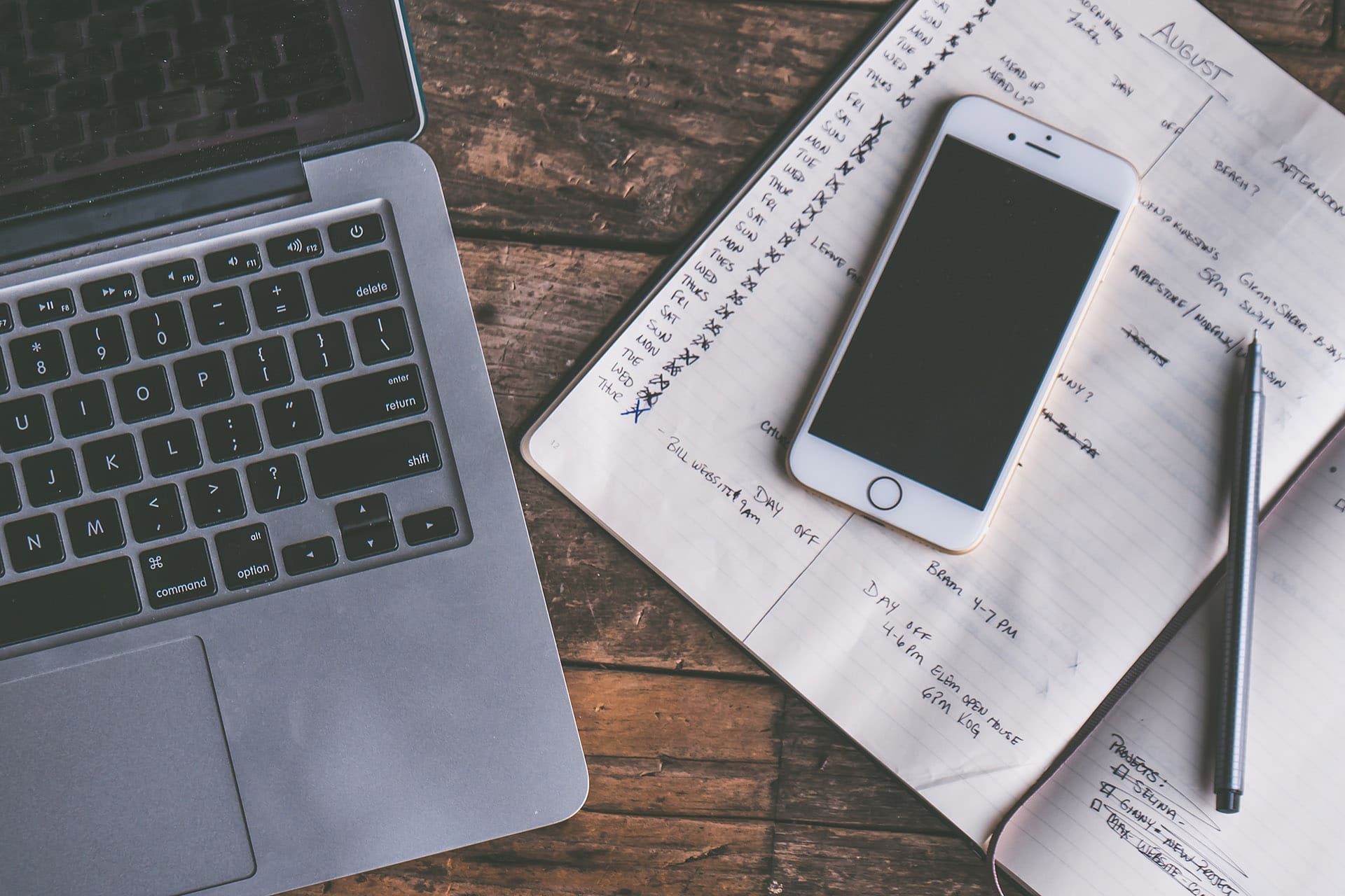 A laptop, iPhone, and pen and notebook sit atop a desk.