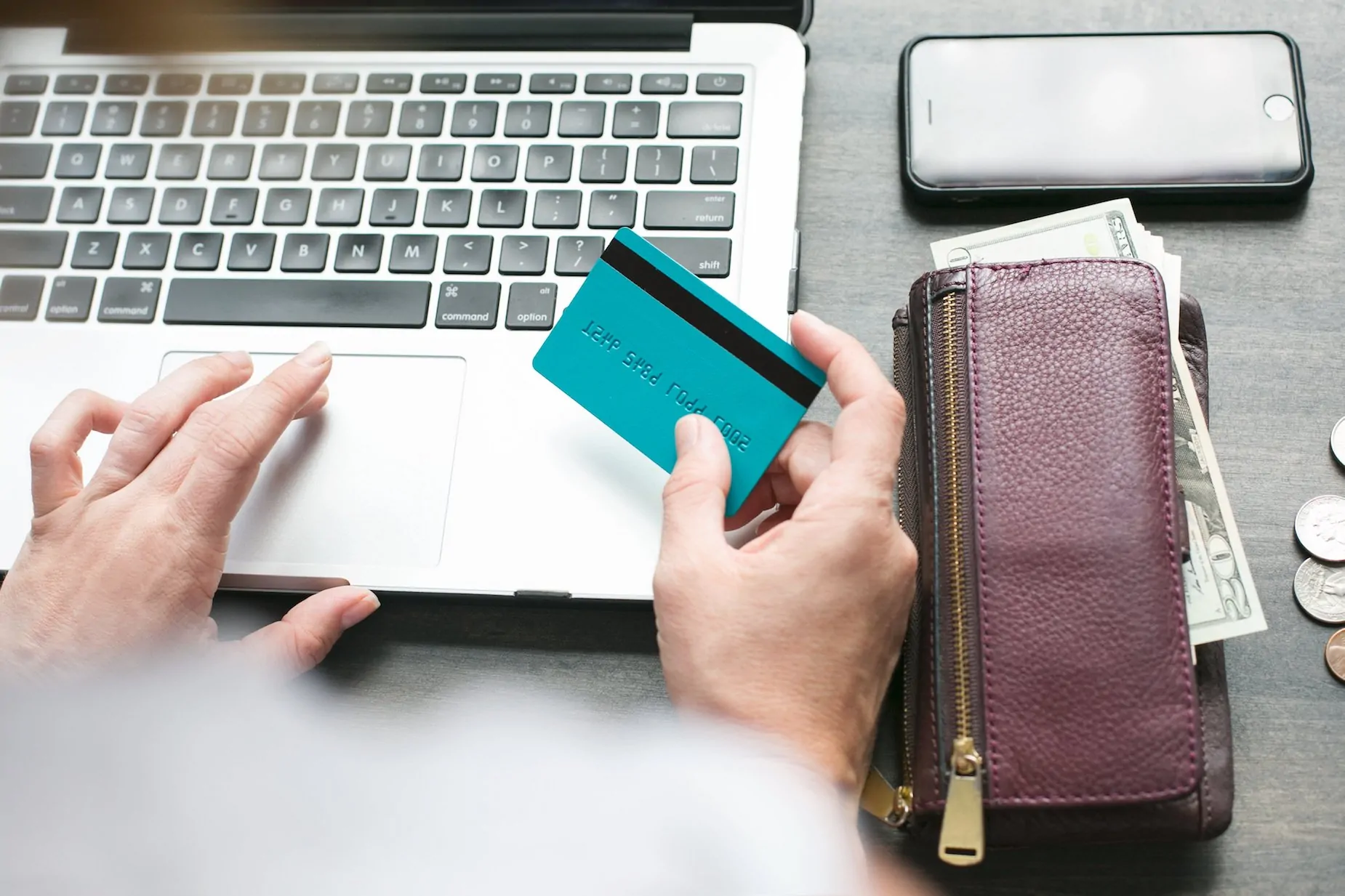 A hand on a laptop, with the other holding a credit card, all beside a wallet stuffed with money.