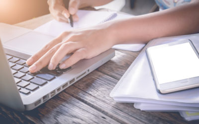 Person with one hand on laptop keyboard and one hand writing on notepad