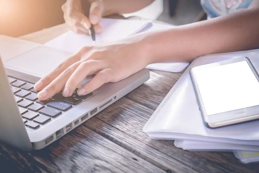 Person with one hand on laptop keyboard and one hand writing on notepad