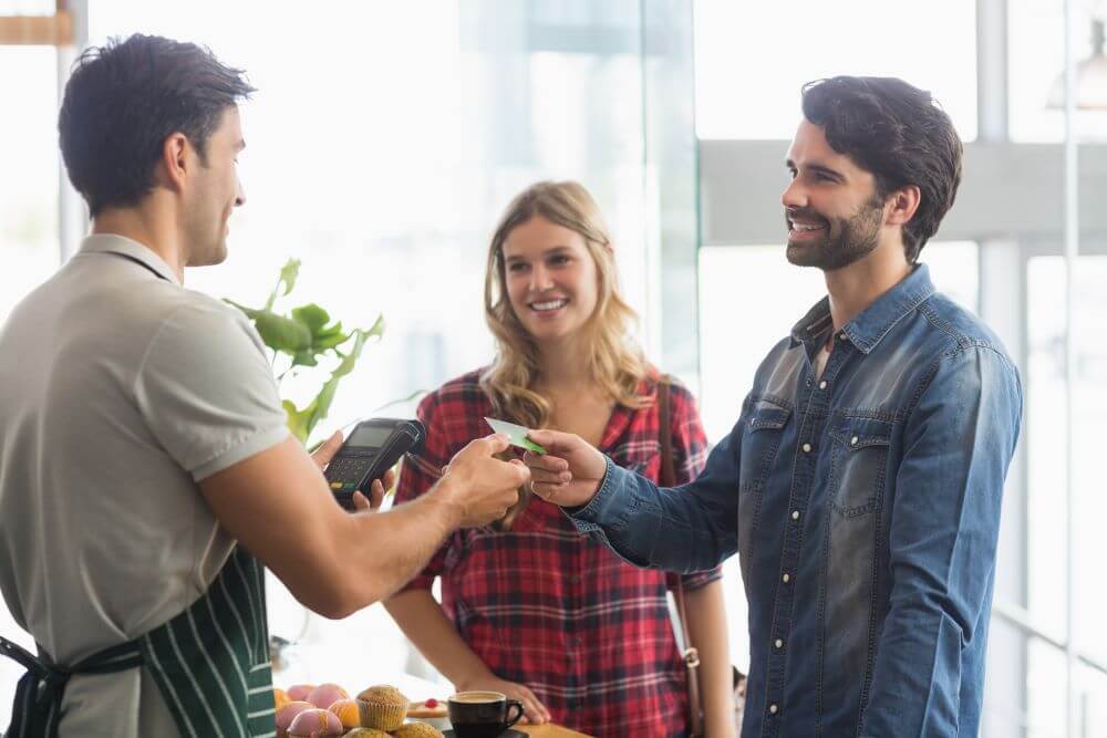 Customer in a blue jean shirt handing his credit card over to a service representative while smiling and standing next to a blonde woman.