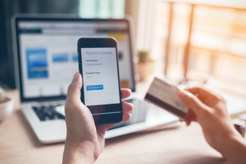 One hand holding smartphone displaying payment details page and other hand holding credit card, with laptop in the background