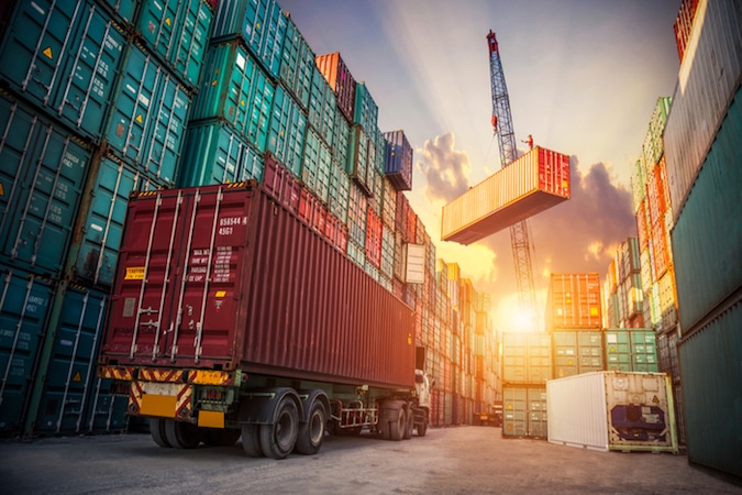 Truck sitting in front of shipping containers with crane lifting one shipping container in the background