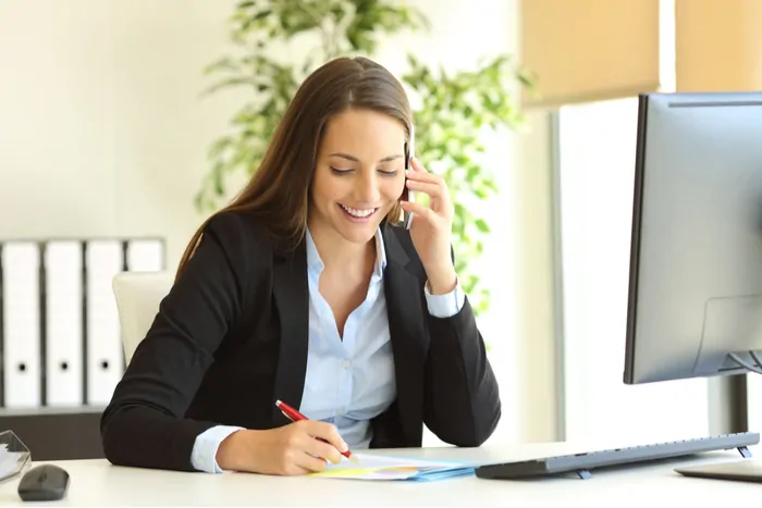 Happy businesswomen talking on cell phone and taking notes at a desk in an office