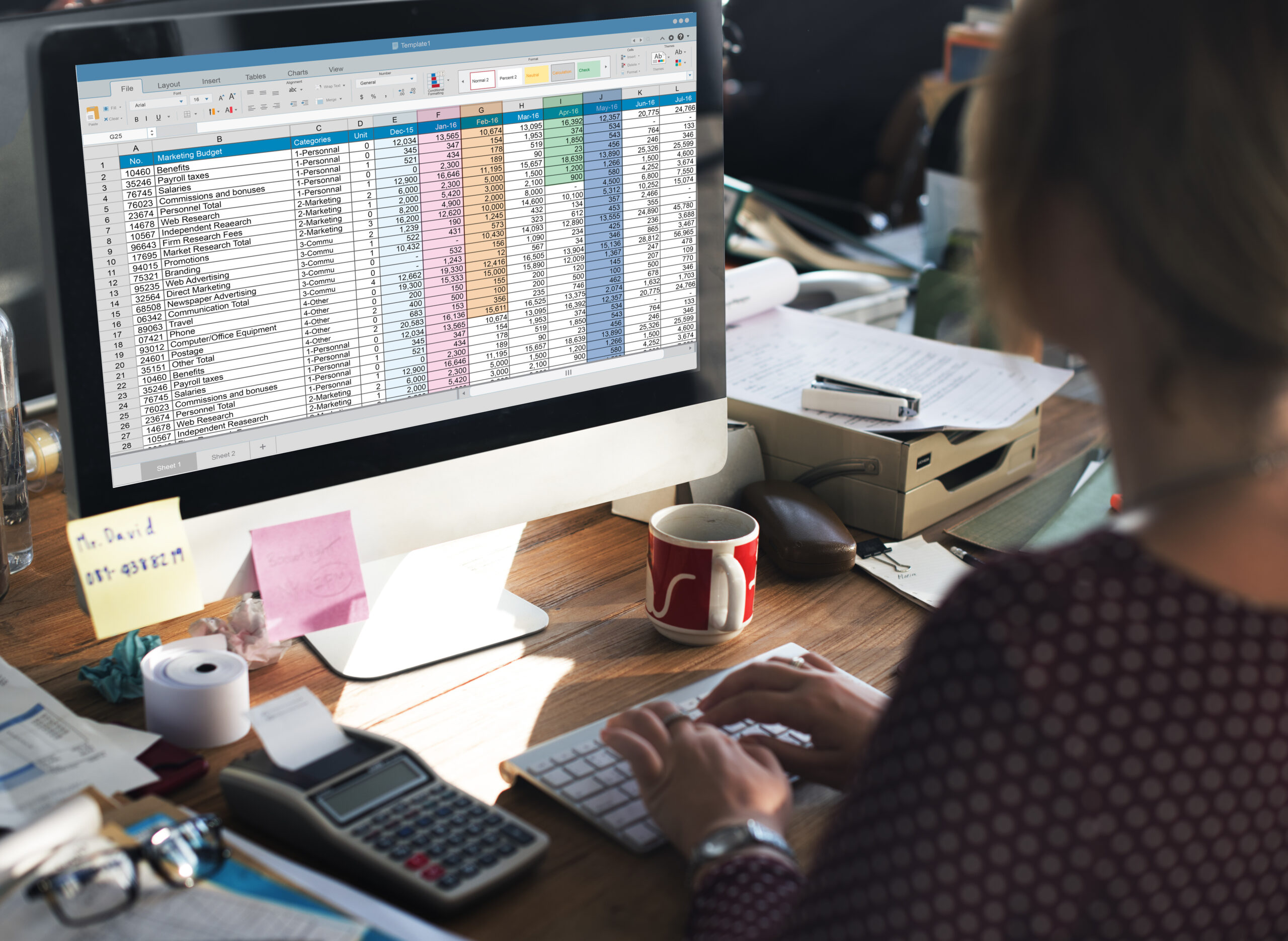 Woman working in a cluttered office using Excel on a Mac Desktop.