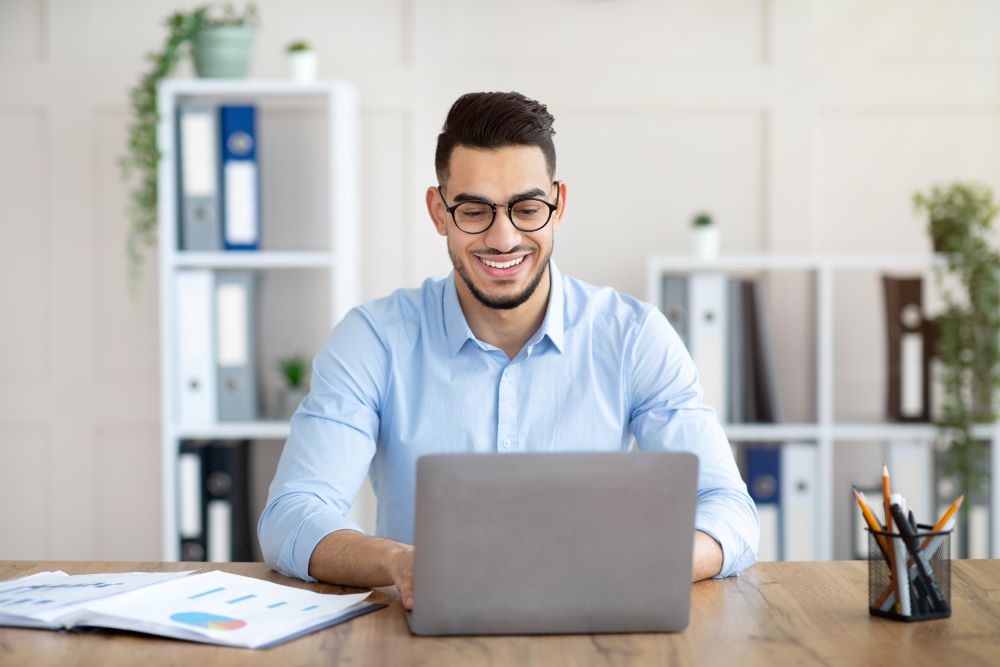 a confident, smiling young man sitting at his office desk and using a laptop