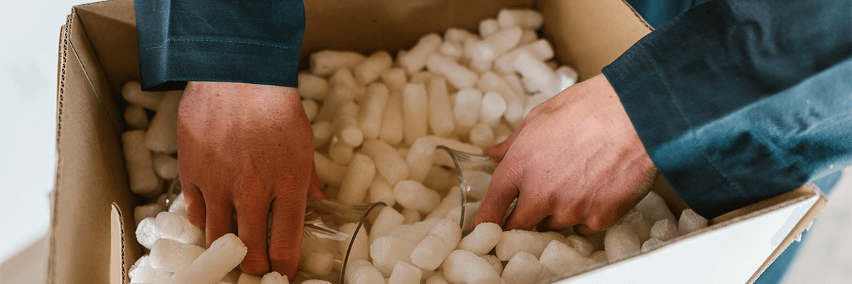 Hands of a person packing glasses into a box filled with packing peanuts.