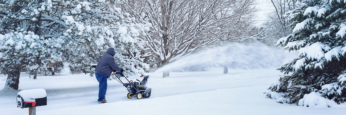 Person pushing a snowblower on a sidewalk surrounded by trees.