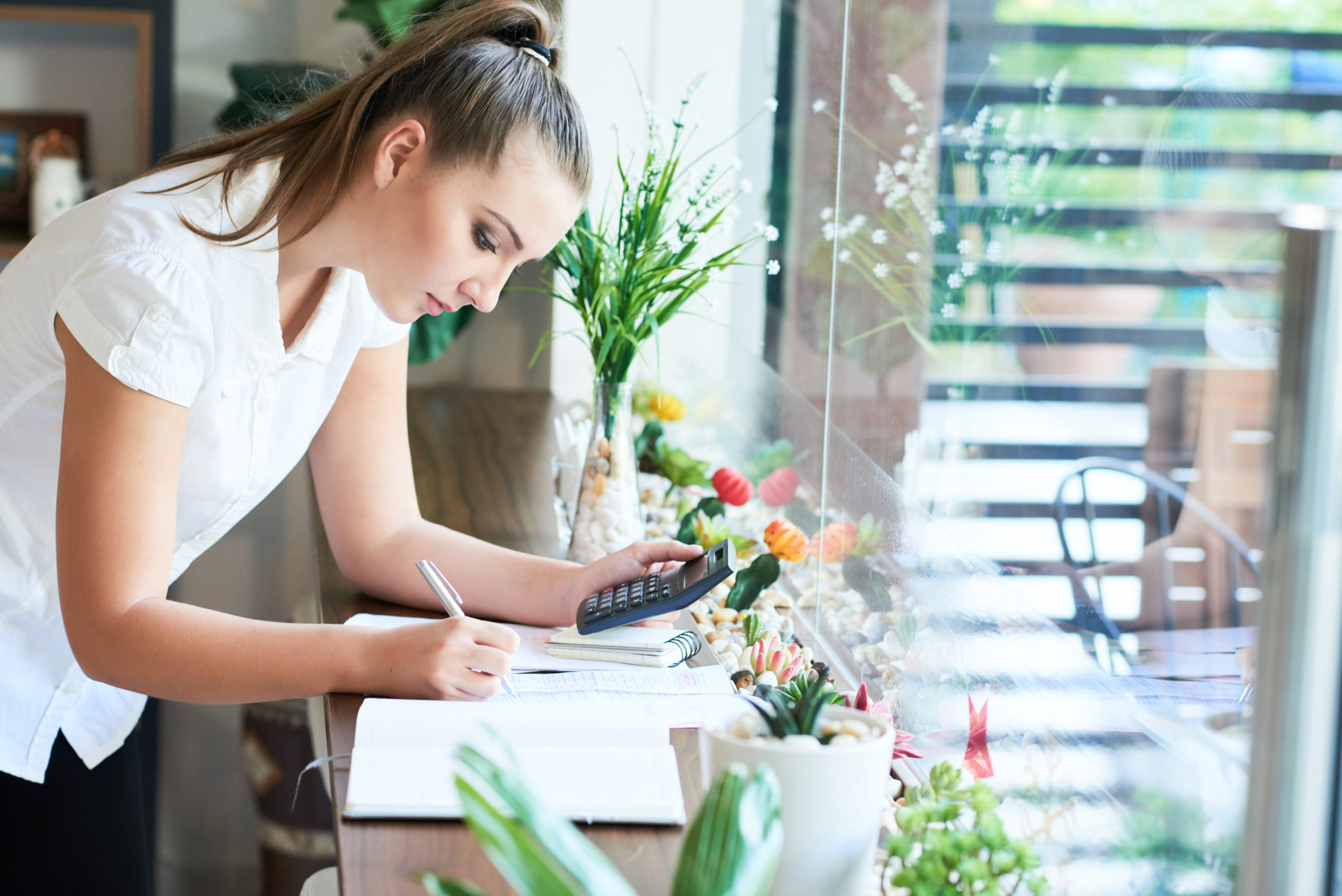 Side view of young formal woman calculating and writing on paper at table near window