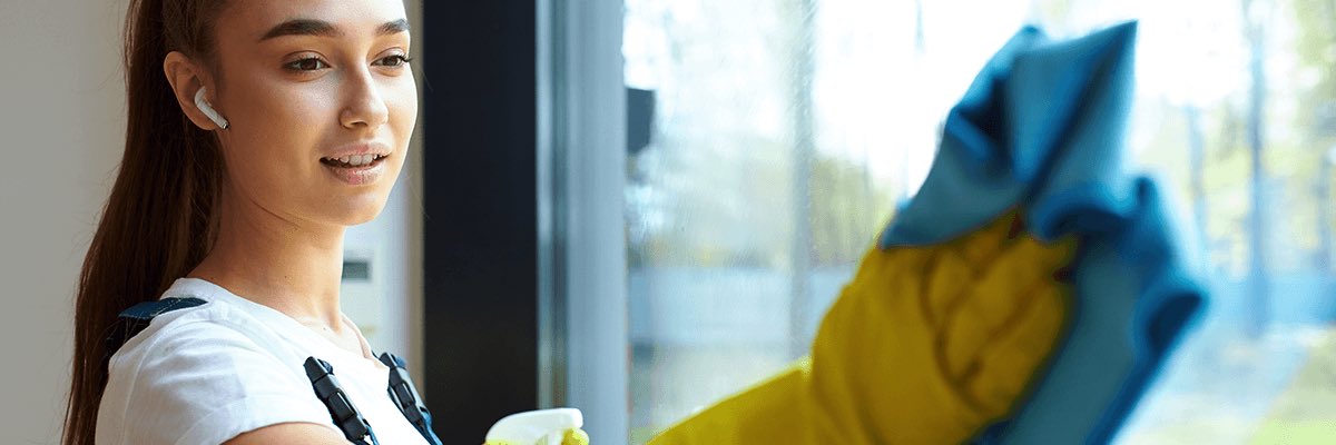 Young woman in uniform, yellow rubber gloves holding blue rag for washing glass, in headphones