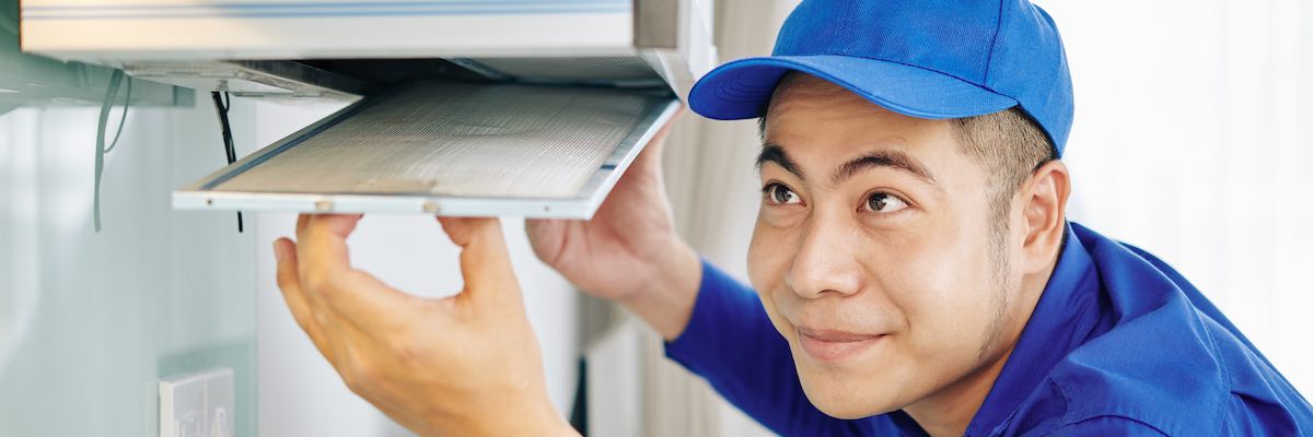 Smiling Asian worker installing cooker hood above stove in kitchen of client