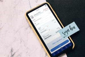 This image shows a smartphone resting atop a file folder, with a blue sticker glued to the screen with the writing "Sign here!".