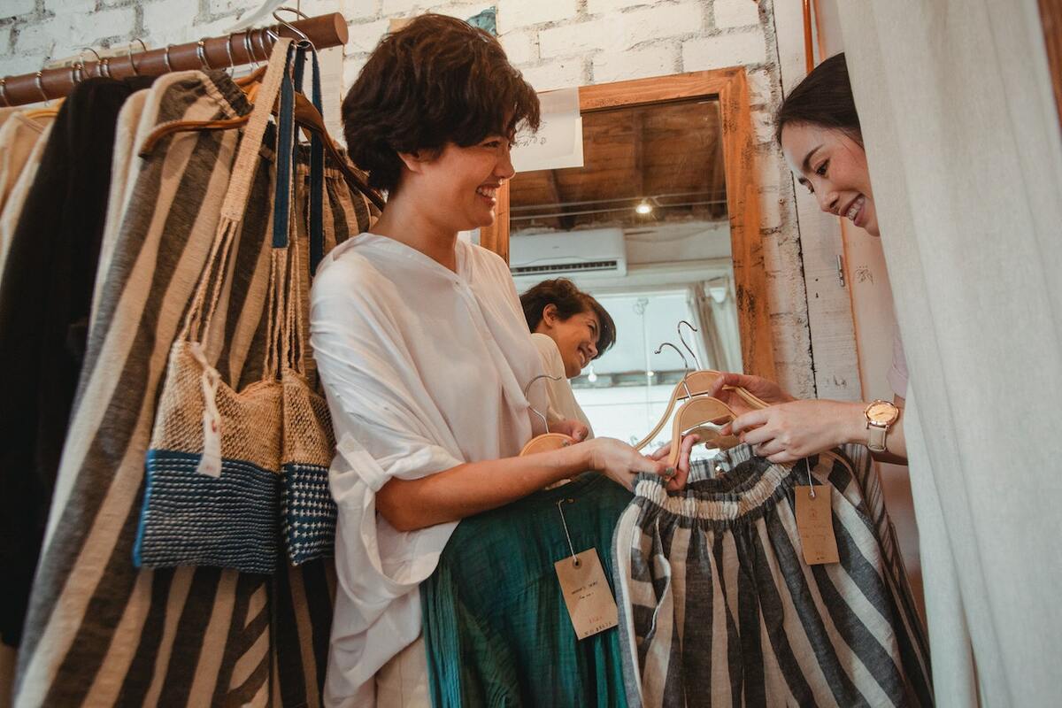 This image features a lady handing a customer clothing pieces to try in the fitting room of a clothing store. Both women are smiling while interacting.