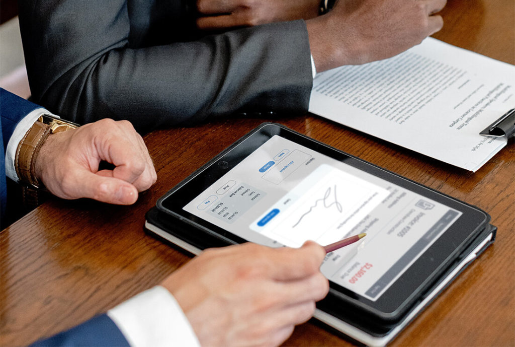 This image shows two people sitting together at a desk. There are documents on the table as well as a tablet device, which is showing a signature capture pop up.