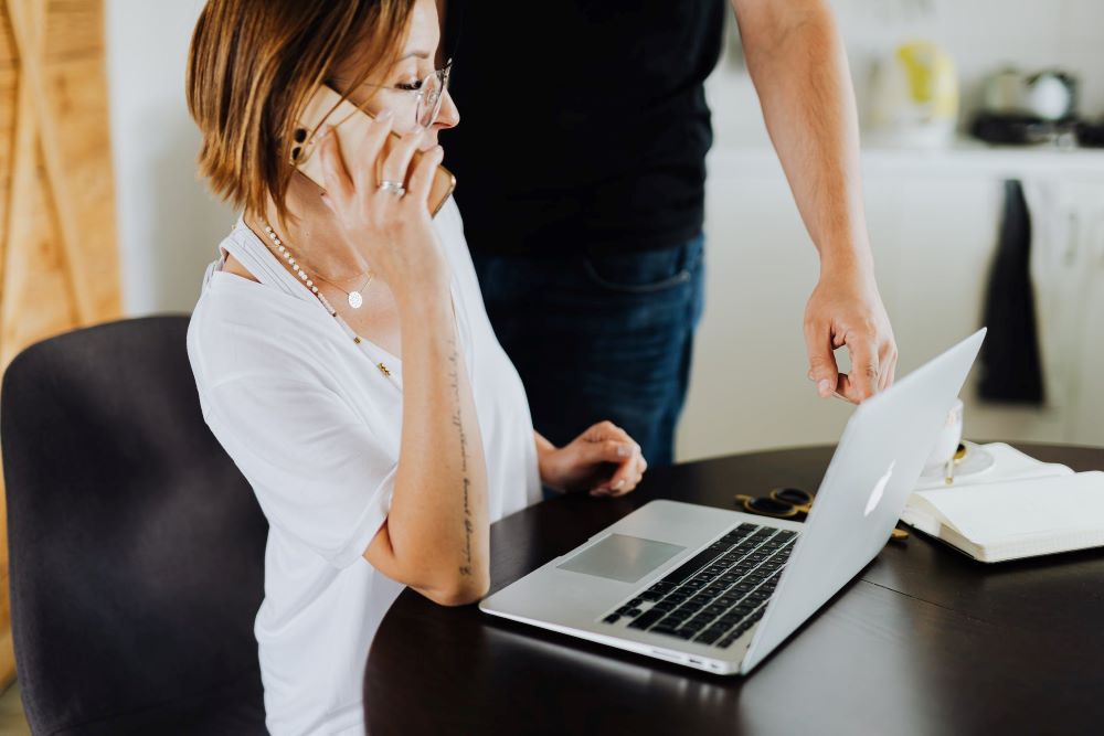 a woman sitting at a desk with a laptop in front of her, while she speaks on the phone.