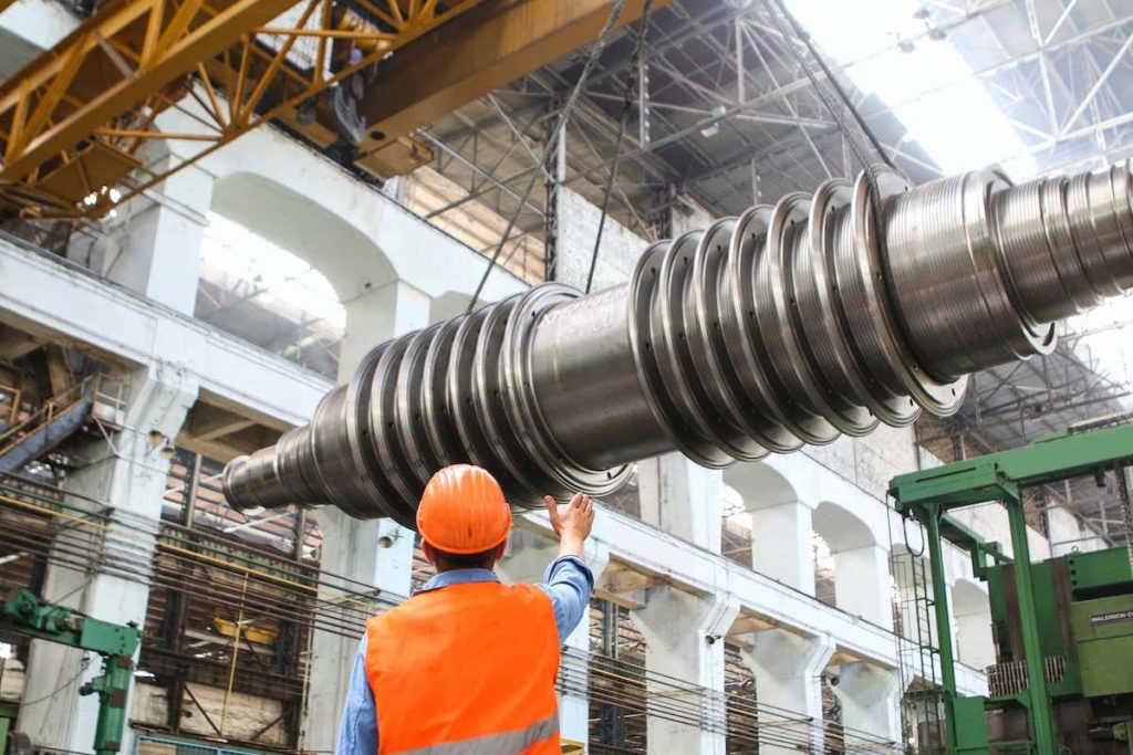 Industrial site with man guiding some large metal...thing. It has the shape of a shock absorber, but it's obviously not that because it's metal. I don't know what it is. Sorry.