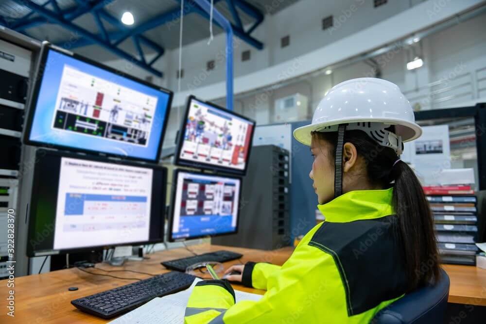 a woman working in a helmet and construction gear, working in an office behind computer screens.