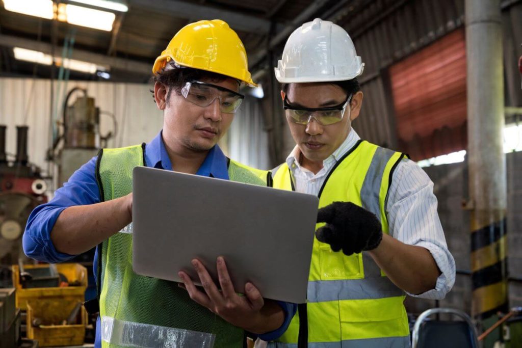 two men wearing protective helmets and neon vests, looking at a tablet and discussing something.