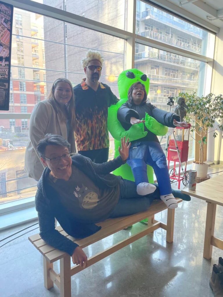 Three Method employees in Halloween costumes and face paint standing in front of a large window and smiling, while one man is laying on a desk, grinning.