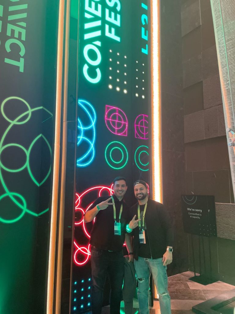 Two young men wearing lanyards and standing in front of a neon wall at the QuickBooks Connect event. 