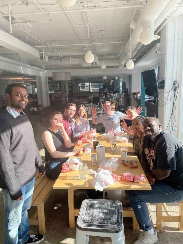 Method team members sitting together at a long table and enjoying lunch, while looking at the camera and smiling
