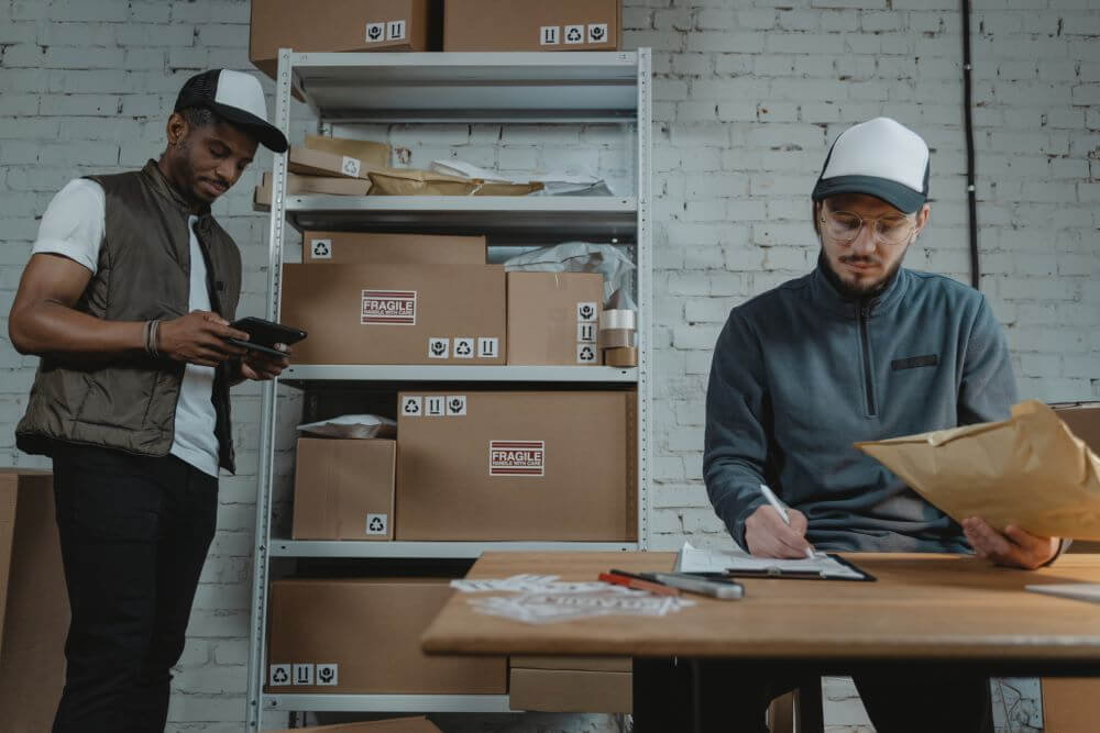 man in a warehouse, sitting at a desk signing documents, while another man who is typing on a phone, stands next to him