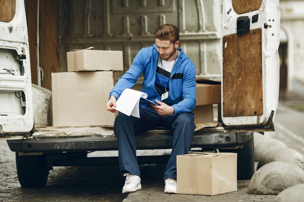 a young man in a blue sweater, looking at a document while sitting at the back of a an open van trunk