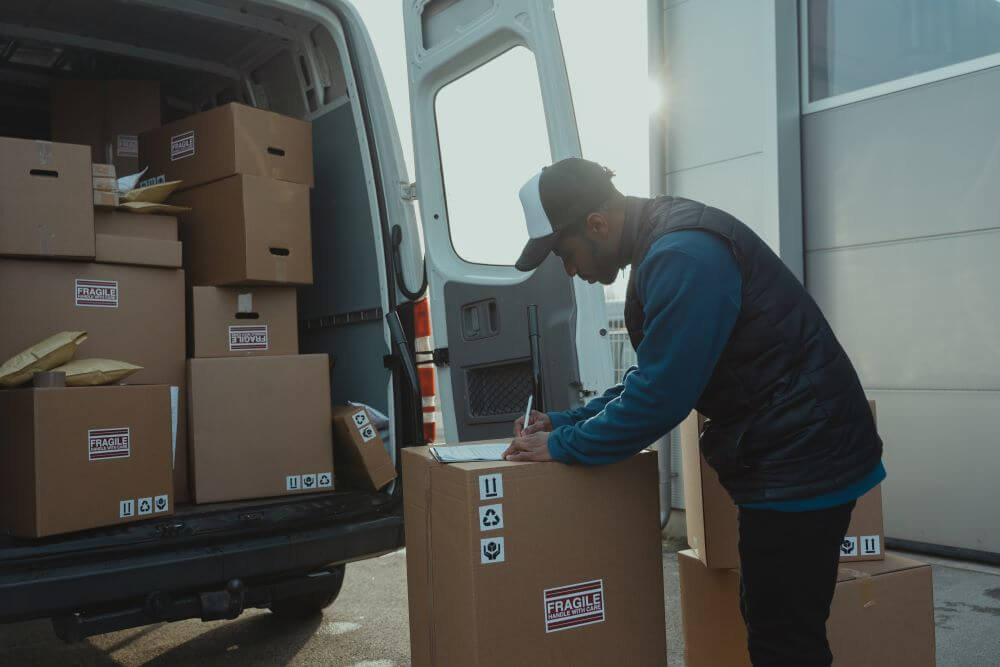 man writing on top of a carton box in front of a van that is loaded with boxes.