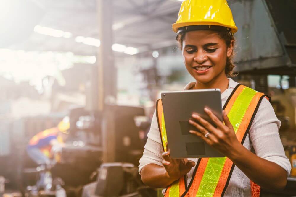 woman in a construction helmet and vest looking at a tablet and smiling.
