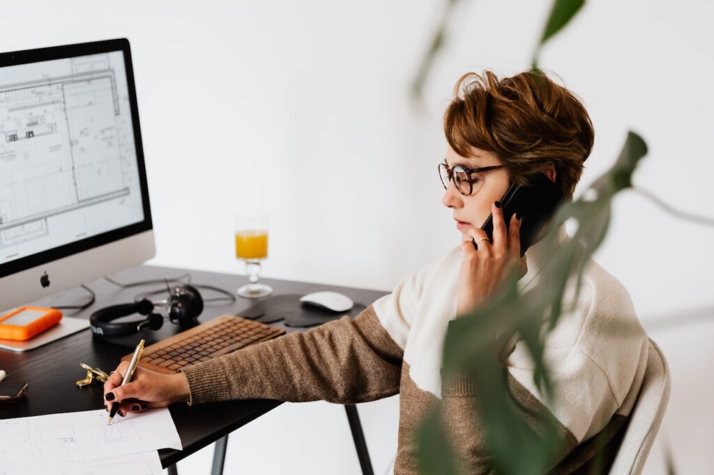 This image shows a businesswoman sat at her desk in front of a computer screen while talking on the phone and taking notes on paper.