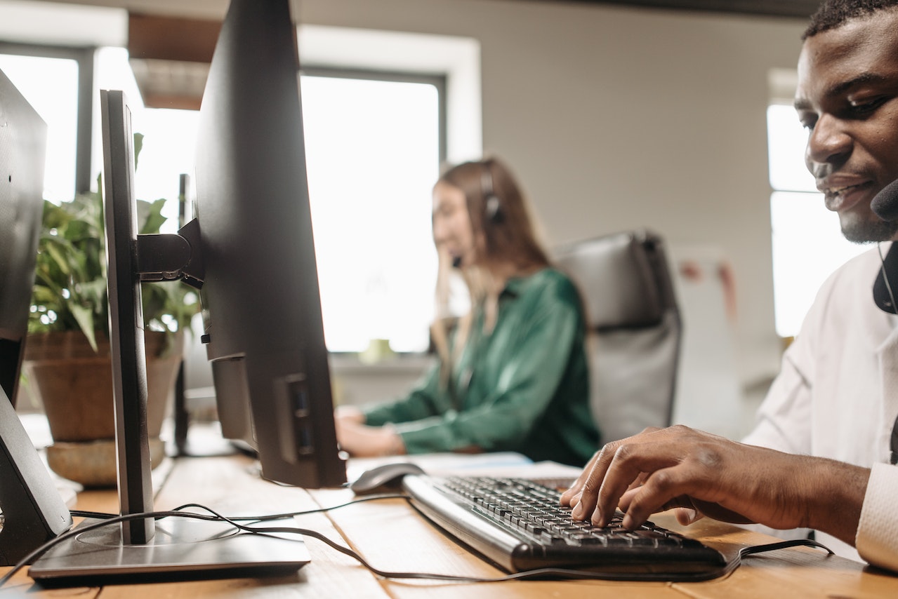 This image shows two office workers sat at a desk working on their computer while talking into headsets.