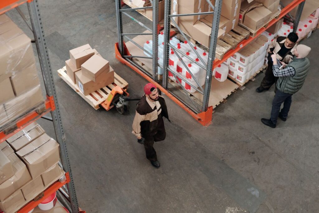 Two men take inventory of a warehouse shelf while another pulls a wagon.