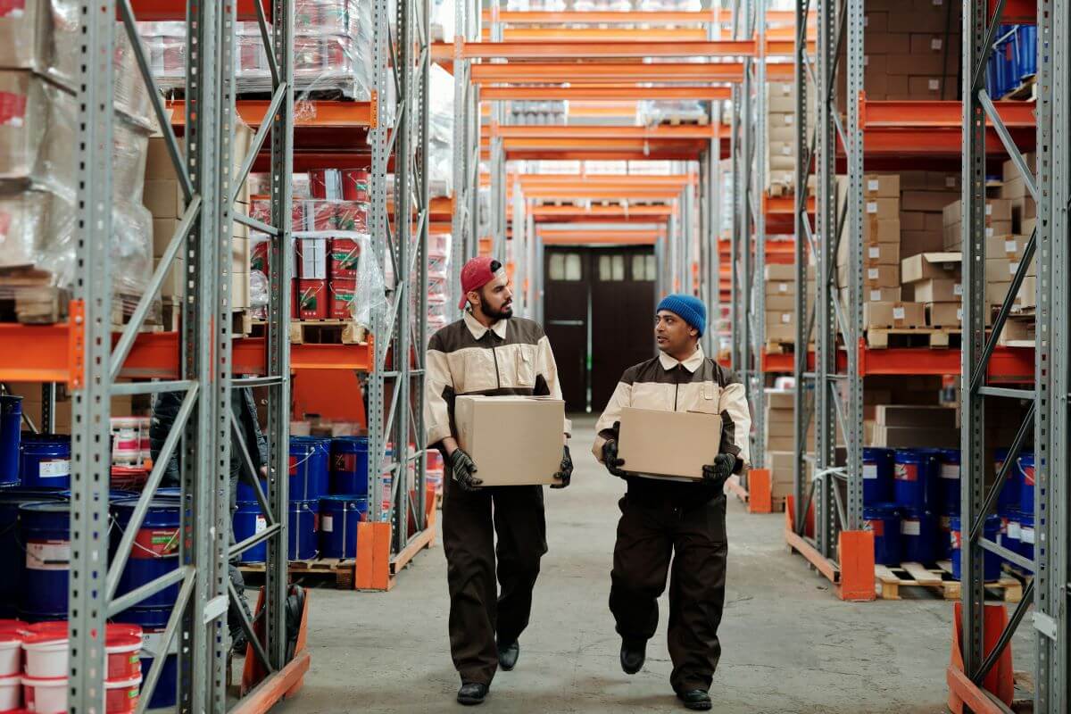 Two men talking and carrying boxes in a warehouse.