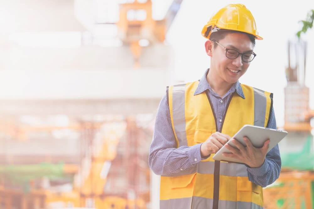 Construction contractor in a yellow vest and helmet looking at a tablet and smiling