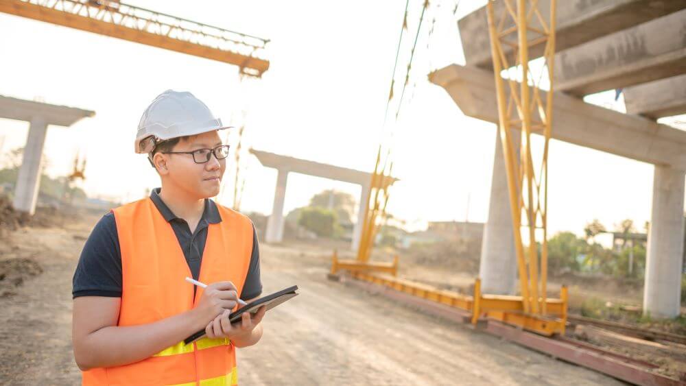 Construction contractor wearing an orange vest and white helmet while holding a tablet and smiling.