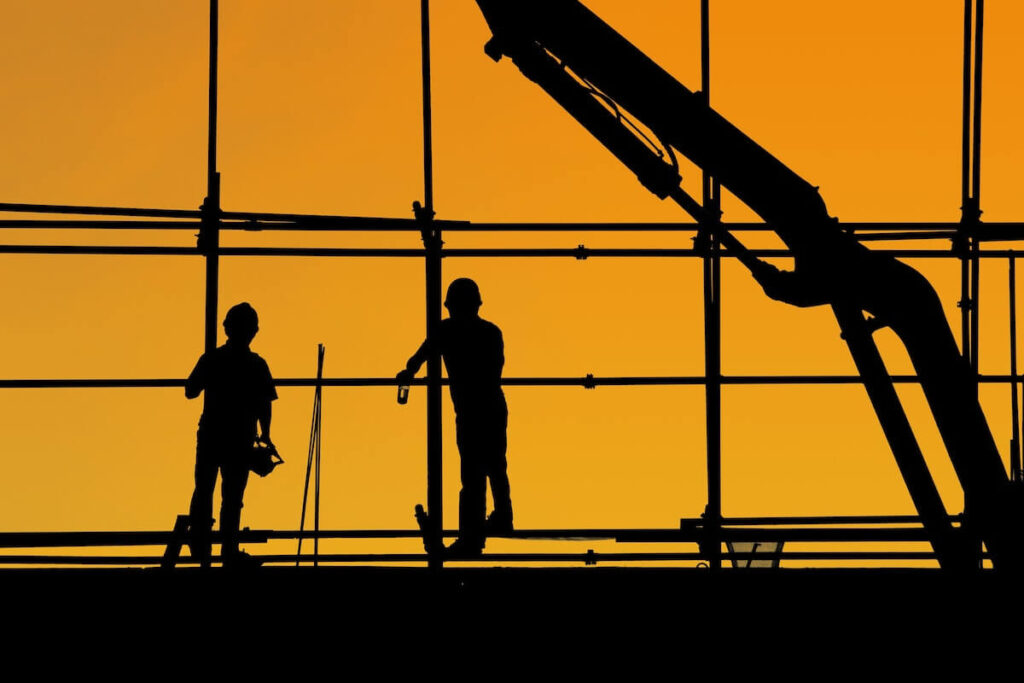 Silhouette of two workers and a crane on a construction site against an orange sky.