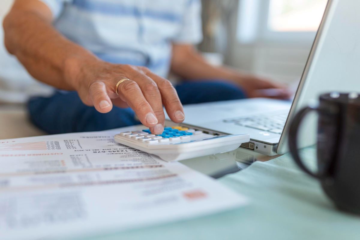 Image shows a man on his laptop while using a calculator that lays over a stack of invoices.