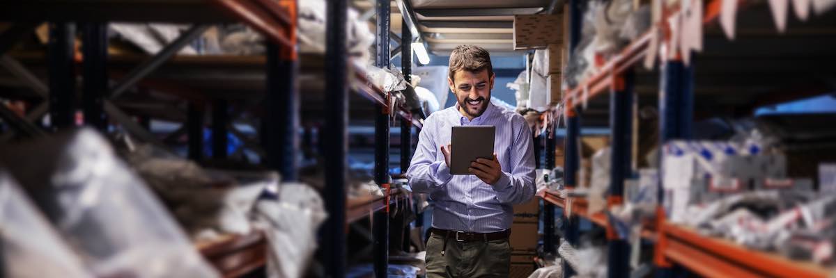 Full length of bearded supervisor walking trough storage of export firm, checking on goods and using tablet.