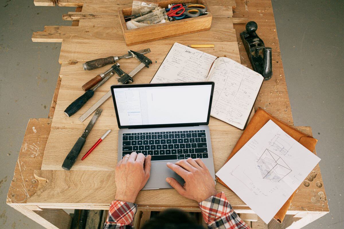 A POV of someone on their laptop, which is on a workspace busy with tools and schematics.