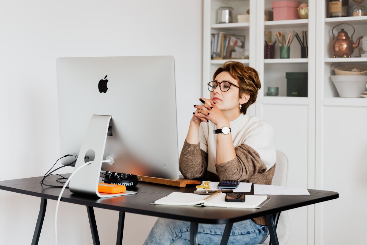 A woman sitting with her hands interlocked, focused on a Mac desktop computer.