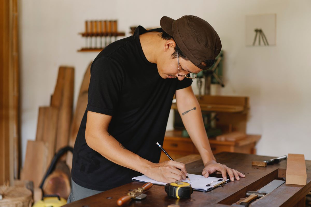 A man writing on a clipboard in a workshop with a tape measure and planks of wood around him.