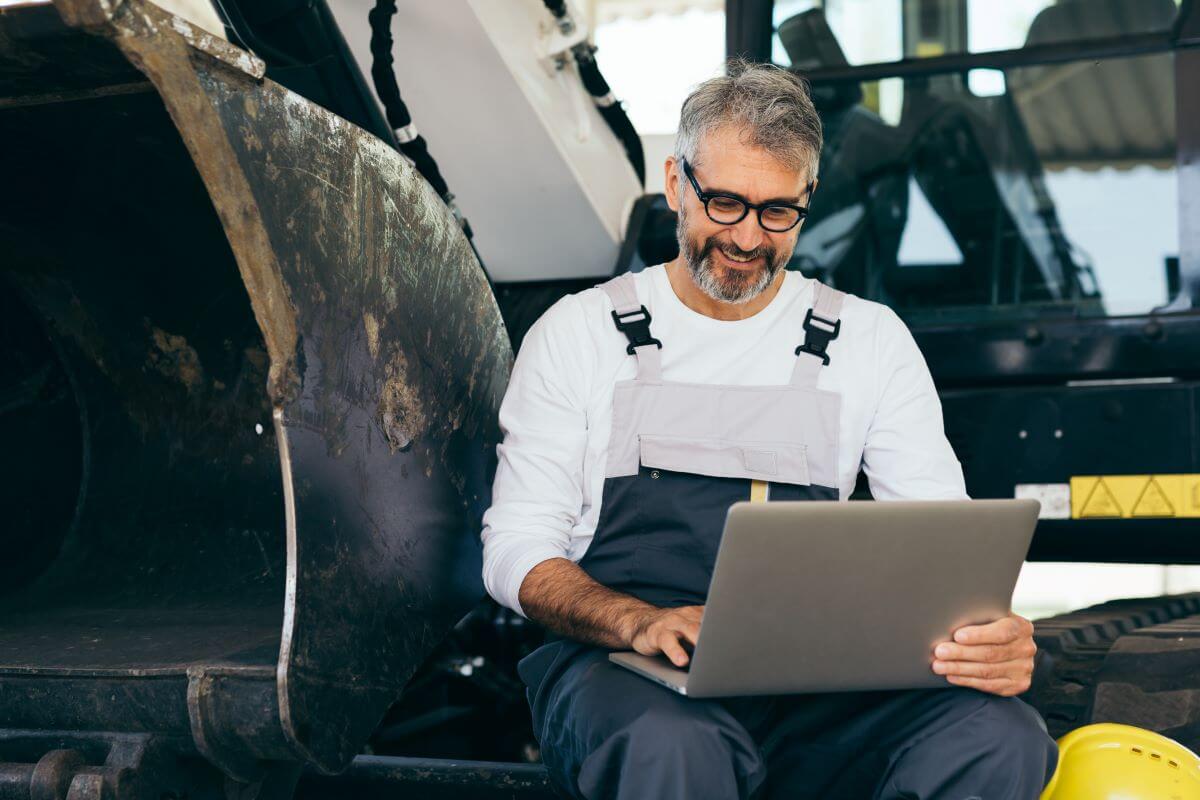 A worker sitting on machinery using a laptop and smiling.