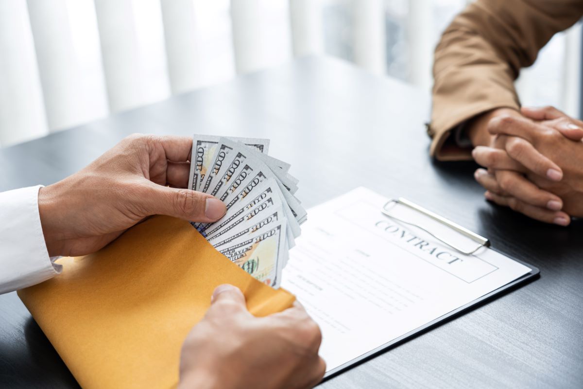 A close-up of two people at a table with a contract on it, and one person is holding an envelope with several US hundred dollar bills.