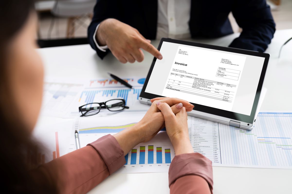 A close-up of a man pointing to an invoice on a tablet; we are looking over the shoulder of a woman reading the tablet with her hands together.