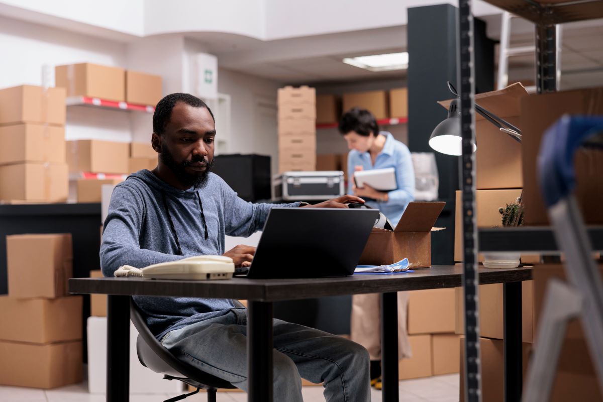 A man in a warehouse full of cardboard boxes sitting at a laptop.