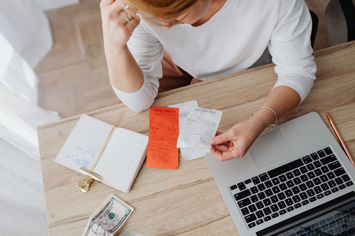 An overhead shot of a woman looking at receipts on a desk. There's a laptop and stack of bills on the desk.