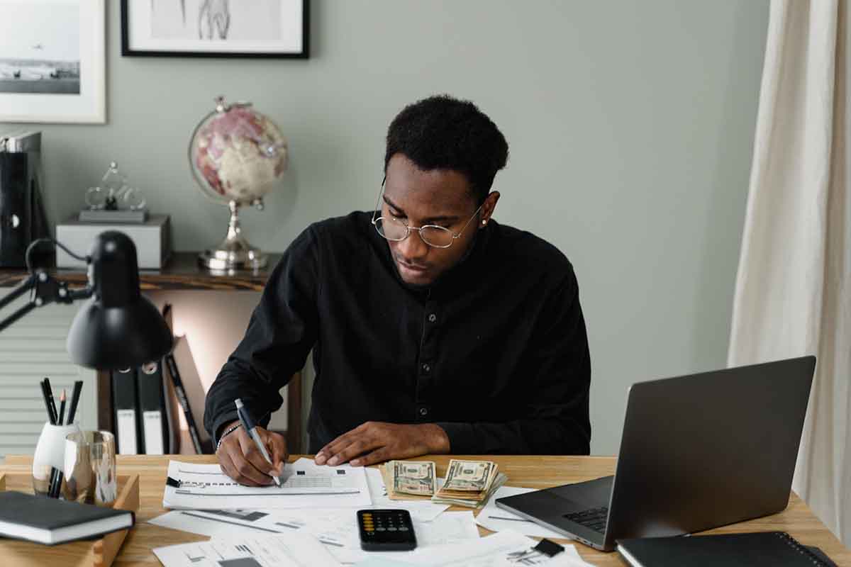 man sitting and writing at a home office with cash, laptop, and a calculator on the desk