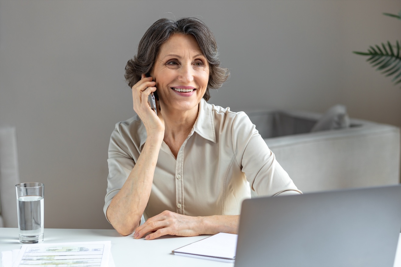 Beautiful senior woman working talking on mobile phone sitting at the workplace with computer laptop in modern office, online remote work, looking away and smiles friendly