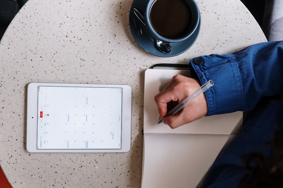 Person writing on a notebook and tablet in front of them on table